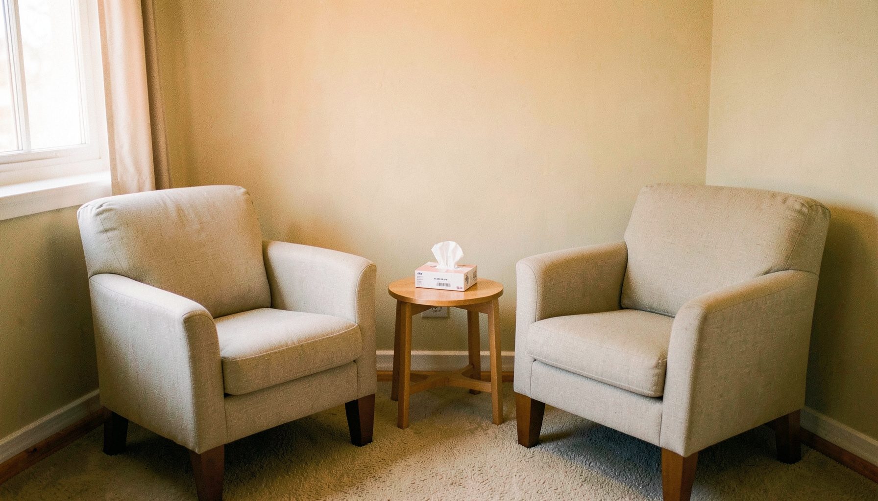 A quiet, softly lit therapy room with two comfortable armchairs facing each other, a small side table with a box of tissues, and warm neutral tones in cream and muted sage